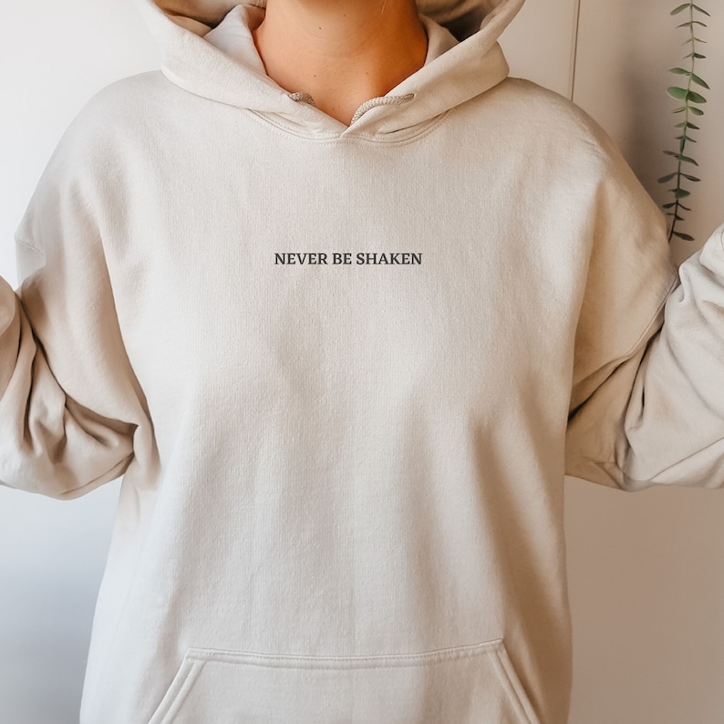 Puede incluir: Una sudadera con capucha beige claro con un mensaje bordado en negro que dice "NEVER BE SHAKEN".
