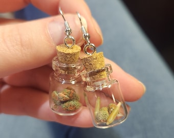 May include: A pair of novelty earrings featuring small glass bottles with cork stoppers. One bottle contains green and orange buds, the other has similar plant matter. The earrings have silver-colored fish hook ear wires.