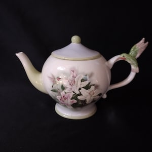 May include: A white ceramic teapot with a green hummingbird on the handle. The teapot is decorated with pink and white flowers.