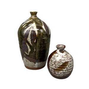 Könnte beinhalten: Zwei Keramikvasen mit rustikaler Ästhetik. Die größere Vase ist hoch und zylindrisch, mit braunen und weißen abstrakten Mustern. Die kleinere Vase ist rund, mit einer ähnlichen Farbpalette und Textur. Beide haben eine glasierte Oberfläche.