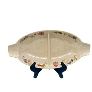 Puede incluir: Fuente para servir dividida de color crema con acabado moteado. La fuente ovalada presenta un diseño caprichoso de setas en rojo, amarillo y gris. Tiene dos asas y está sostenida por un soporte negro, perfecta para servir aperitivos.