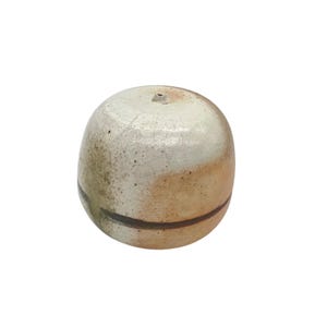 Può includere: Piccolo oggetto rotondo in ceramica, beige chiaro e marrone. Presenta un piccolo foro sulla parte superiore e una fascia marrone scuro al centro. L'oggetto ha un aspetto rustico e artigianale.
