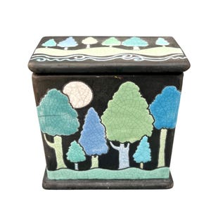 May include: A black ceramic box with a lid, featuring a landscape design. The box is adorned with stylized trees in shades of green and blue, a white moon, and a green base. The lid has a row of trees and a wavy line design.