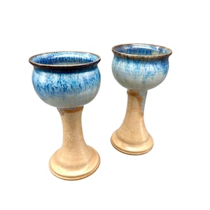 Pode incluir: Dois cálices de cerâmica feitos à mão. As taças são de um azul vibrante com um padrão salpicado, que transita para um azul mais claro perto da base. As hastes e bases são de uma cor bege quente, criando um contraste marcante.