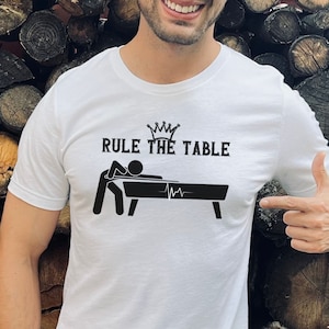May include: White t-shirt with a black graphic of a person playing pool with the text "RULE THE TABLE" and a crown above the text.