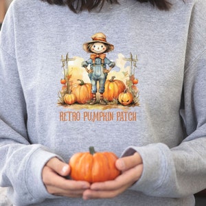 May include: A light gray sweatshirt featuring a watercolor illustration of a scarecrow in a hat and overalls standing in a pumpkin patch. The words "Retro Pumpkin Patch" are printed below the image. An orange pumpkin is held in the hands.