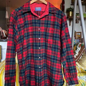 May include: A long-sleeve button-down shirt featuring a red, dark green, and navy blue plaid pattern. The shirt has a pointed collar, a single chest pocket, and a red quilted lining visible at the inner neckline with a Pendleton brand label.