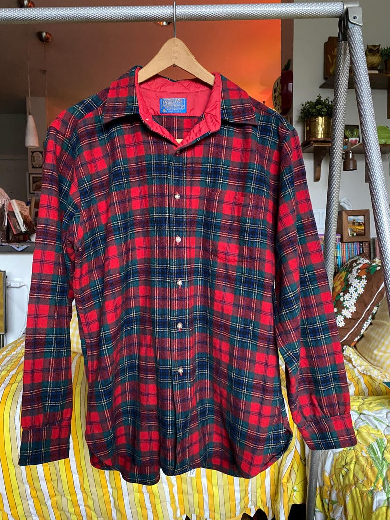 May include: A long-sleeve button-down shirt featuring a red, dark green, and navy blue plaid pattern. The shirt has a pointed collar, a single chest pocket, and a red quilted lining visible at the inner neckline with a Pendleton brand label.