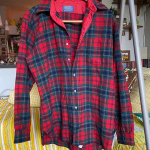 May include: A long-sleeve button-down shirt featuring a red, dark green, and navy blue plaid pattern. The shirt has a pointed collar, a single chest pocket, and a red interior lining along the collar and button placket. A Pendleton label is visible.