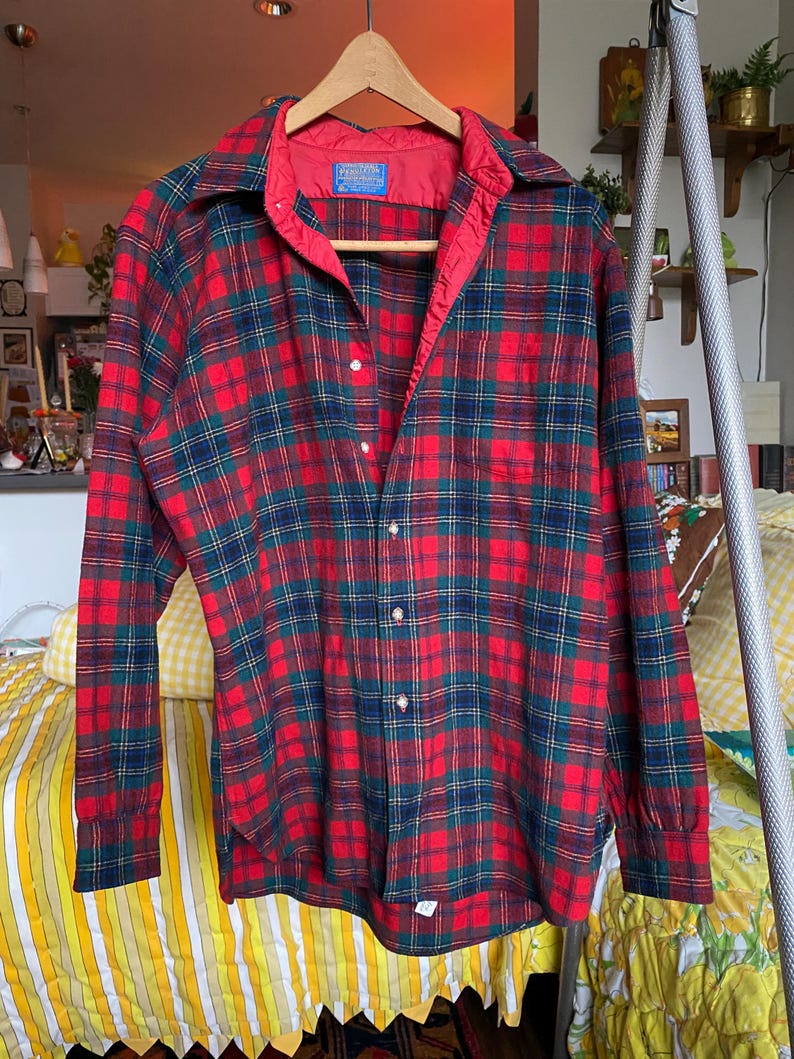 May include: A long-sleeve button-down shirt featuring a red, dark green, and navy blue plaid pattern. The shirt has a pointed collar, a single chest pocket, and a red interior lining along the collar and button placket. A Pendleton label is visible.
