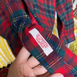 May include: A long-sleeved shirt featuring a red, green, and blue plaid tartan pattern. A white fabric label is sewn onto the interior cuff, printed with the red text AUTHENTIC MAC LEAN TARTAN. The garment has a red lining visible at the cuff edge.
