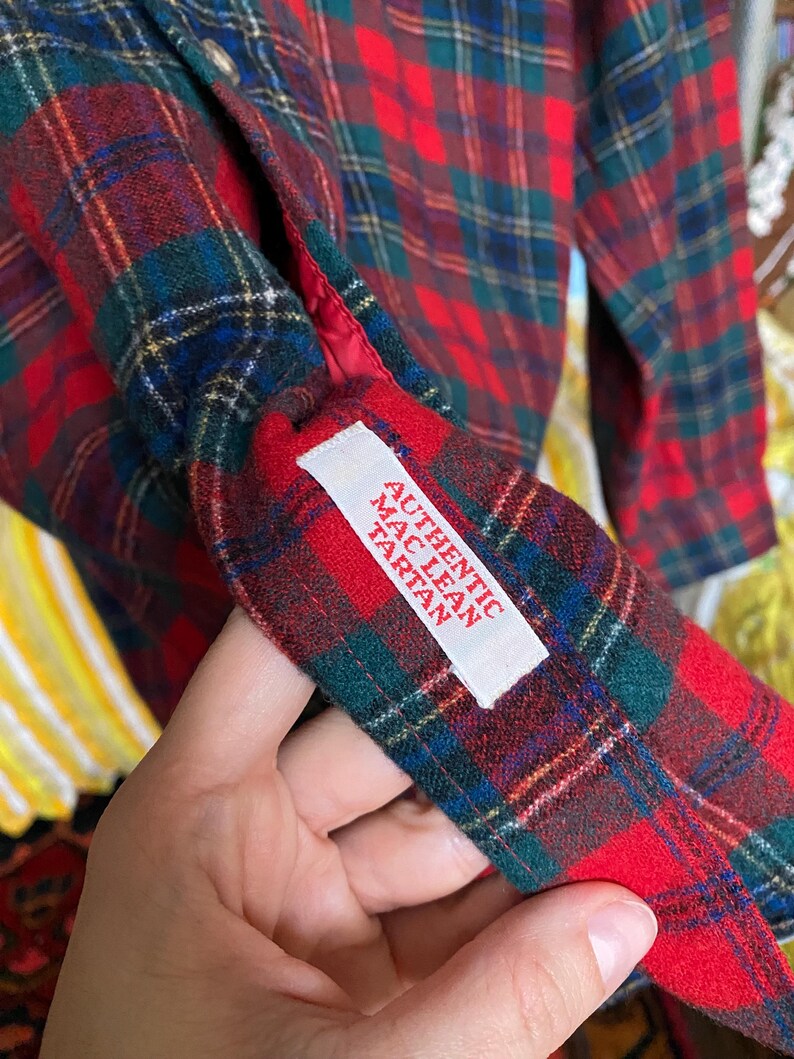 May include: A long-sleeved shirt featuring a red, green, and blue plaid tartan pattern. A white fabric label is sewn onto the interior cuff, printed with the red text AUTHENTIC MAC LEAN TARTAN. The garment has a red lining visible at the cuff edge.