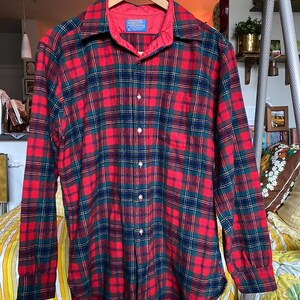 May include: A long-sleeve button-down shirt featuring a red, dark green, and navy blue plaid pattern. The shirt has a pointed collar, a single chest pocket, and a red interior lining at the neck. A blue Pendleton brand label is visible at the collar.