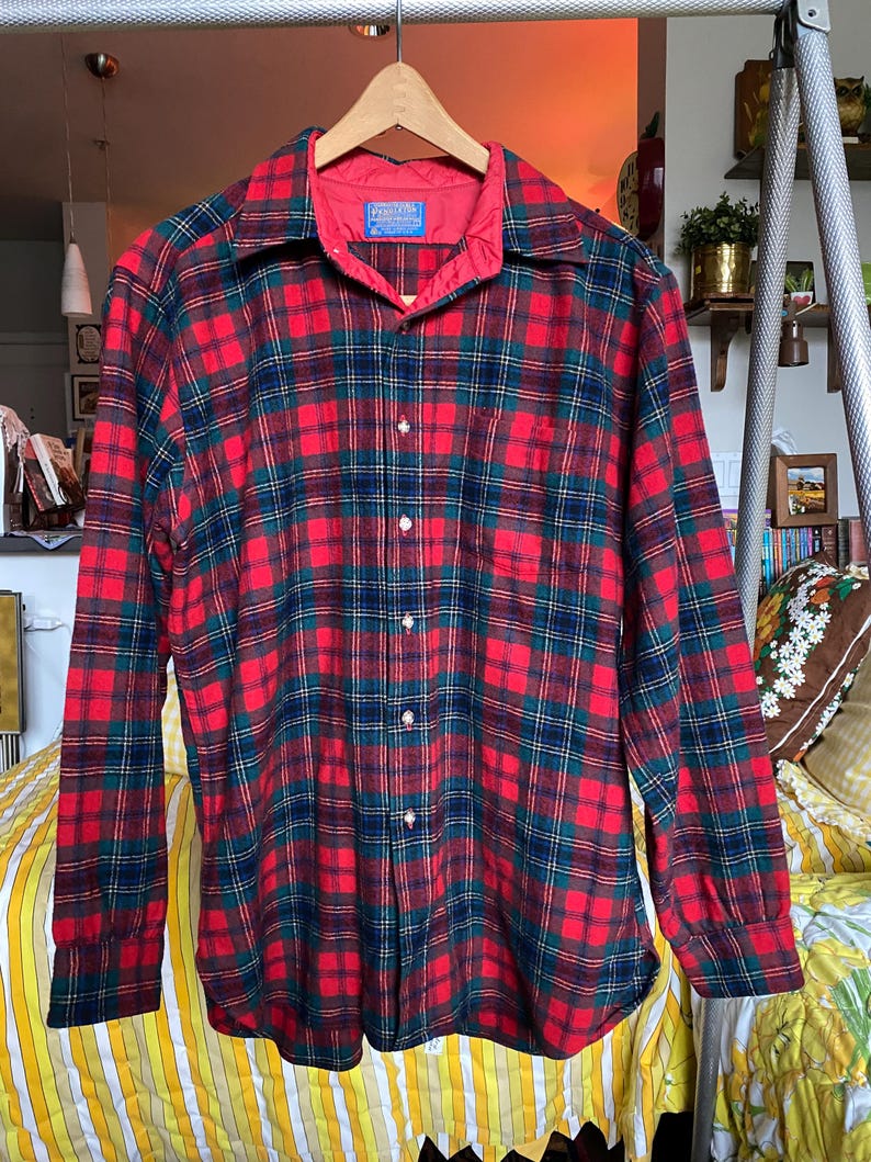 May include: A long-sleeve button-down shirt featuring a red, dark green, and navy blue plaid pattern. The shirt has a pointed collar, a single chest pocket, and a red interior lining at the neck. A blue Pendleton brand label is visible at the collar.