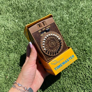 May include: A vintage brown Signature Timemaster multiple program timer in its original yellow and brown box. The timer has a circular dial with numbers and a switch for on, off, and auto settings. The box has the text "Timemaster" and "multiple program time".