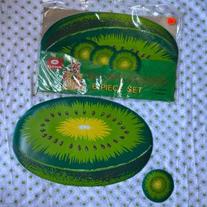 May include: A set of kiwi-shaped placemats and coasters. The placemats are green with a yellow center and brown seeds, resembling a kiwi fruit slice. The packaging reads "8 PIECE SET".