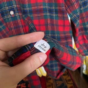 May include: A long-sleeve button-down shirt featuring a red, green, and blue plaid pattern. A white interior tag is visible, clearly printed with the text 'dry clean' in black lettering. The shirt has a light-colored button on the front placket.