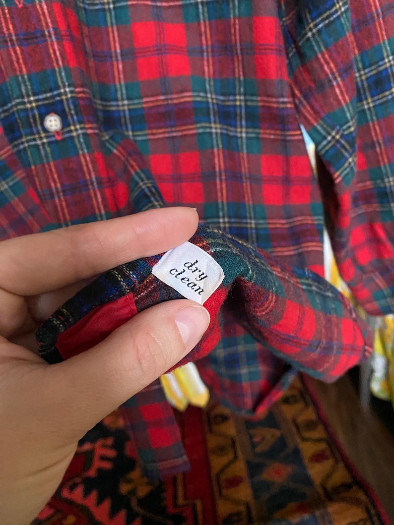 May include: A long-sleeve button-down shirt featuring a red, green, and blue plaid pattern. A white interior tag is visible, clearly printed with the text 'dry clean' in black lettering. The shirt has a light-colored button on the front placket.