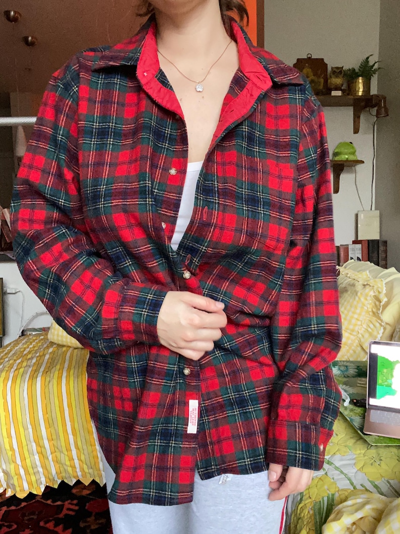 May include: A long-sleeved button-down shirt featuring a red, dark green, and navy blue plaid pattern. The shirt has a classic collar, a front button closure, and a small rectangular white label with red text near the bottom hem.