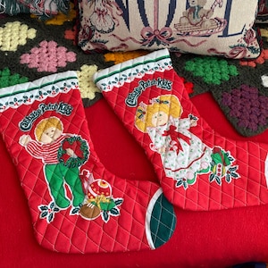 May include: Two red quilted Christmas stockings with Cabbage Patch Kids designs. One stocking features a boy holding a wreath, the other a girl in a dress. Both have green and white accents.