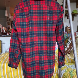 May include: A long-sleeved button-down shirt featuring a vibrant plaid pattern in shades of red, dark green, and navy blue. The shirt has a classic collar, a buttoned front placket, and cuffs with single buttons, displayed on a wooden hanger.