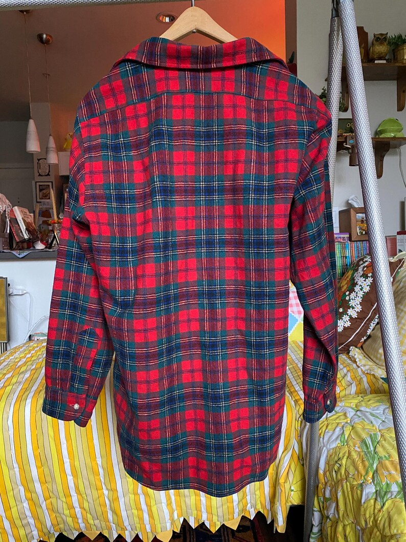 May include: A long-sleeved button-down shirt featuring a vibrant plaid pattern in shades of red, dark green, and navy blue. The shirt has a classic collar, a buttoned front placket, and cuffs with single buttons, displayed on a wooden hanger.