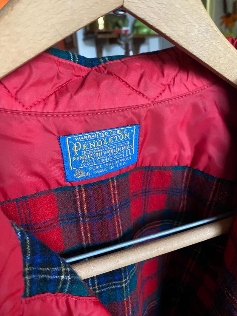 May include: A Pendleton jacket with a red quilted lining and a red, green, and blue plaid wool exterior. A blue rectangular label is stitched to the interior, reading: Warranted to be a Pendleton, Pendleton Woolen Mills, Portland, Oregon, 100% Virgin Wool, L, Pure Virgin Wool, Made in U.S.A.