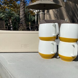 May include: Four white ceramic mugs with a mustard yellow base and a rectangular beige container are displayed on a concrete surface. The mugs have a rounded shape with a handle. The background includes outdoor furniture and foliage.