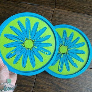 May include: Two circular coasters featuring a bright lime green background with a large, stylized blue daisy flower design. Each coaster has a thick, raised blue border and a smooth, flat surface. The floral pattern is outlined in black ink.
