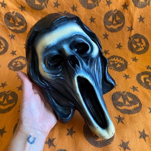 May include: A plastic ghost-themed mask featuring a distorted, elongated face with hollow black eye sockets and a wide, open mouth. The mask has a glossy finish with a gradient of off-white and deep black shading, creating a dramatic, high-contrast appearance.