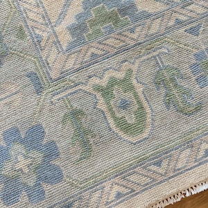 Op de afbeelding: Close-up van een beige tapijt met een gedetailleerd patroon. Het ontwerp bevat blauwe en groene bloemen en geometrische vormen. Het tapijt heeft een franje langs de rand, wat textuur en detail toevoegt.