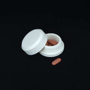 May include: A white plastic pill container with a lid. The container is open and there are several brown pills inside.