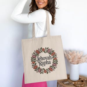 May include: Beige tote bag featuring a floral wreath design with the words "Jehovah Rapha" in the center. The wreath includes red, pink, and green elements. The bag has long handles for carrying.