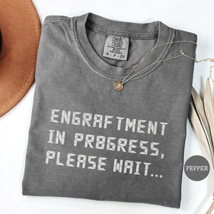 May include: A gray Comfort Colors t-shirt with the text "ENGRAFTMENT IN PROGRESS, PLEASE WAIT..." in white. A gold necklace with a pendant is draped on the shirt. A brown hat is in the upper left corner. The shirt is labeled "PEPPER".