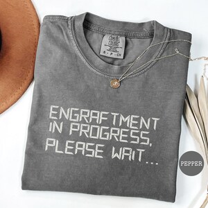 May include: A gray t-shirt with the text "ENGRAFTMENT IN PROGRESS, PLEASE WAIT..." in white. A gold necklace with a pendant is on the shirt. The shirt is folded on a white surface.