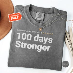May include: Gray t-shirt with the text "TRANSPLANT WARRIOR 100 days Stronger" in white. The shirt has a distressed look and is a comfortable, casual style.