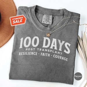 May include: Grey t-shirt with the text "100 Days Post Transplant Resilience - Faith - Courage" printed in white.
