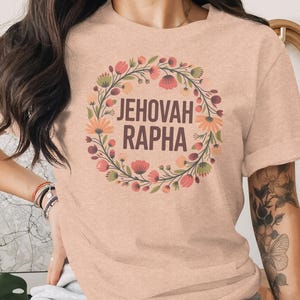 May include: A peach-colored t-shirt featuring the words "JEHOVAH RAPHA" in a stylized font, encircled by a floral wreath in shades of pink, orange, and green. The shirt has short sleeves and a crew neck.
