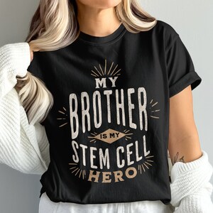 Op de afbeelding: Zwart t-shirt met de tekst "MY BROTHER IS MY STEM CELL HERO" in een distressed, vintage stijl. De tekst is in wit en bruin, met stralende lijnen. Het shirt heeft een ronde hals.