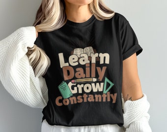Op de afbeelding: Zwart T-shirt met de tekst "Learn Daily Grow Constantly" in bruine en oranje letters. Het ontwerp bevat een boek, een potlood en een liniaal.