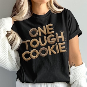 Könnte beinhalten: Schwarzes T-Shirt mit dem Schriftzug "ONE TOUGH COOKIE" in einer Schriftart, die an Kekse erinnert. Die Buchstaben haben ein strukturiertes, gebackenes Aussehen. Die Person trägt ein weißes Strickjäckchen.