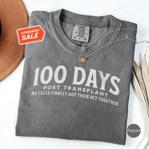 May include: Gray t-shirt with the text "100 Days Post Transplant My Cells Finally Got Their Act Together" in white lettering.