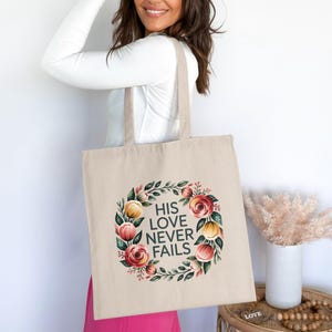 May include: A beige tote bag featuring a floral wreath design and the text "HIS LOVE NEVER FAILS". The wreath includes red, pink, and yellow flowers with green leaves. The bag is rectangular with long handles, suitable for carrying items.
