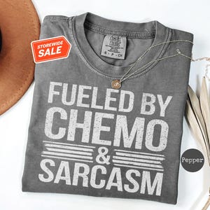 Puede incluir: Una camiseta gris Comfort Colors con el texto "Fueled by Chemo & Sarcasm" en blanco. Una pequeña pegatina de "Storewide Sale" está adjunta. Un collar dorado con un colgante está sobre la camiseta.