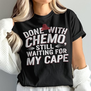 May include: Black t-shirt with white text that reads "Done with chemo, still waiting for my cape".