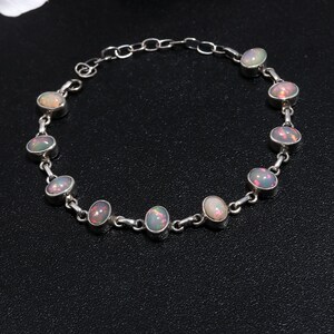 Puede incluir: Un brazalete de plata con piedras de ópalo de forma ovalada. El brazalete tiene un diseño de cadena delicado y presenta un total de nueve piedras de ópalo.