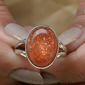 Puede incluir: Un anillo de plata con una piedra preciosa ovalada de color naranja con inclusiones brillantes. La banda del anillo se divide en dos delicadas puntas a cada lado de la piedra preciosa.