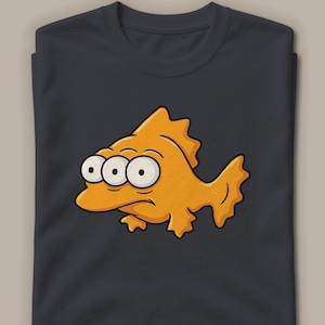 Könnte beinhalten: Ein dunkel anthrazitfarbenes T-Shirt mit einer grafischen Illustration eines dreiaugigen orangefarbenen Fisches. Der Fisch hat einen runden Körper, gezackte Flossen und einen neutralen Ausdruck. Das Motiv ist mittig auf der Brust des T-Shirts platziert.