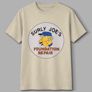 Könnte beinhalten: Beiges T-Shirt mit einem kreisförmigen Aufdruck mit der Aufschrift "SURLY JOE'S FOUNDATION REPAIR". Die Grafik zeigt eine Zeichentrickfigur mit blauem Hut und Zigarre. Das T-Shirt hat kurze Ärmel und einen Rundhalsausschnitt.