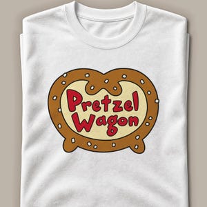 Op de afbeelding: Een wit t-shirt met korte mouwen voorzien van een grote afbeelding van een bruine krakeling met witte zoutkristallen. Binnenin de krakelingvorm staat de tekst "Pretzel Wagon" gedrukt in dikke rode letters op een lichtcrèmekleurige achtergrond.